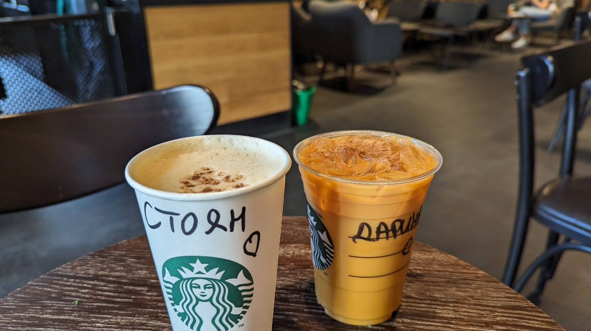 Two Starbucks drinks on a round wooden table: a hot beverage with foam and cinnamon in a paper cup, and an iced orange drink in a plastic cup, both labeled with names written in Cyrillic script. Cafe seating is visible in the background.