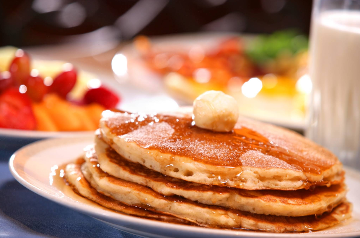 A stack of pancakes topped with a pat of butter and drizzled with syrup sits on a plate, part of a cheap all you can eat breakfast, with a glass of milk and a colorful fruit bowl in the blurred background.