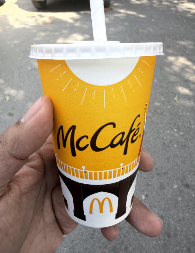 A hand holding a yellow McCafé cup with a white lid and straw, featuring the McCafé logo and a McDonald's arch symbol, outdoors on a sunny day.