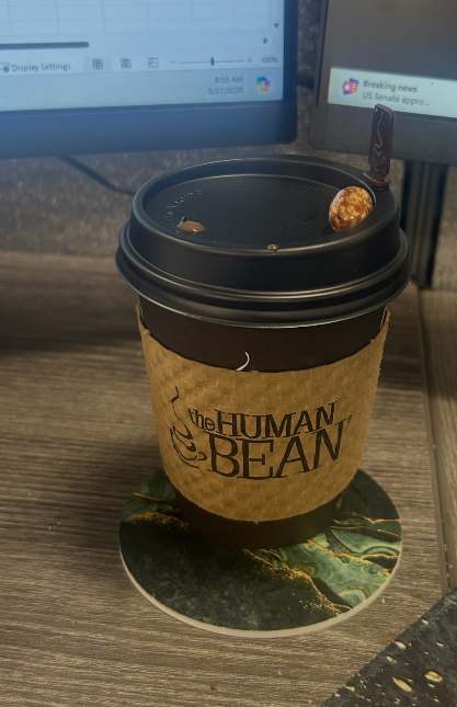 A hot coffee cup with a brown sleeve labeled "the HUMAN BEAN" sits on a colorful coaster in front of two computer monitors, one showing a news alert. A chocolate-covered espresso bean rests on the lid.