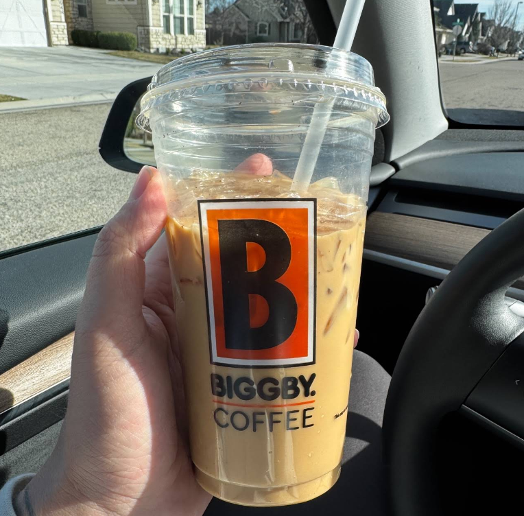 A hand holding a clear plastic cup of iced coffee with a straw, labeled "BIGGBY COFFEE," inside a car with sunlight shining through the window.
