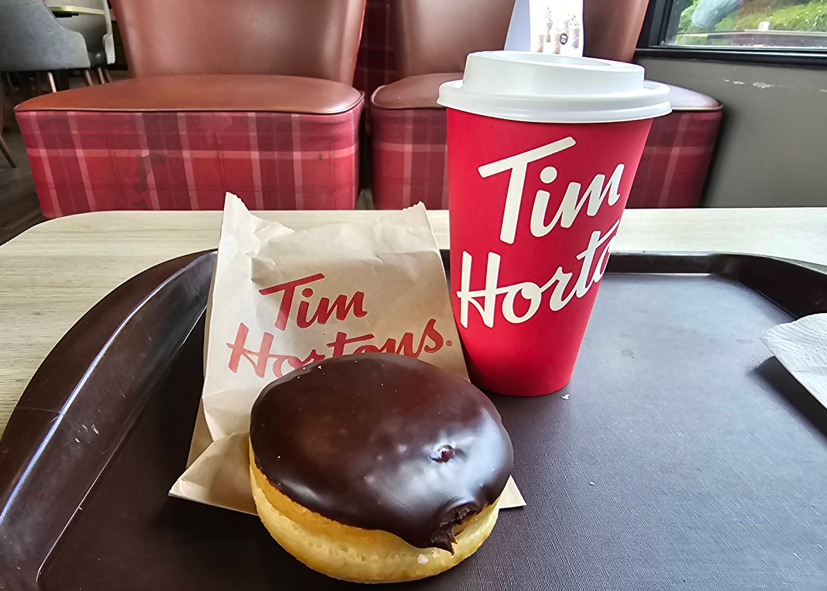 A Tim Hortons coffee cup and a chocolate-glazed donut on a tray, with a paper bag labeled "Tim Hortons" in a cafe setting with red plaid chairs in the background.