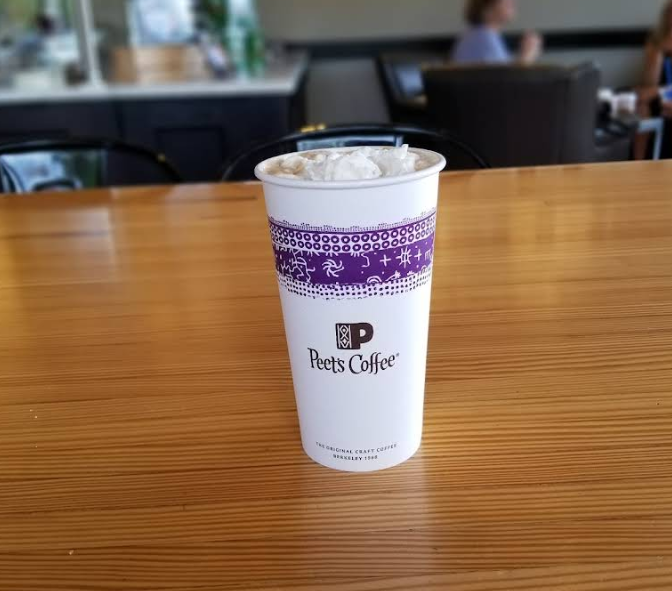A cup of Peet's Coffee topped with whipped cream sits on a wooden table in a café, with blurred seating and people in the background.