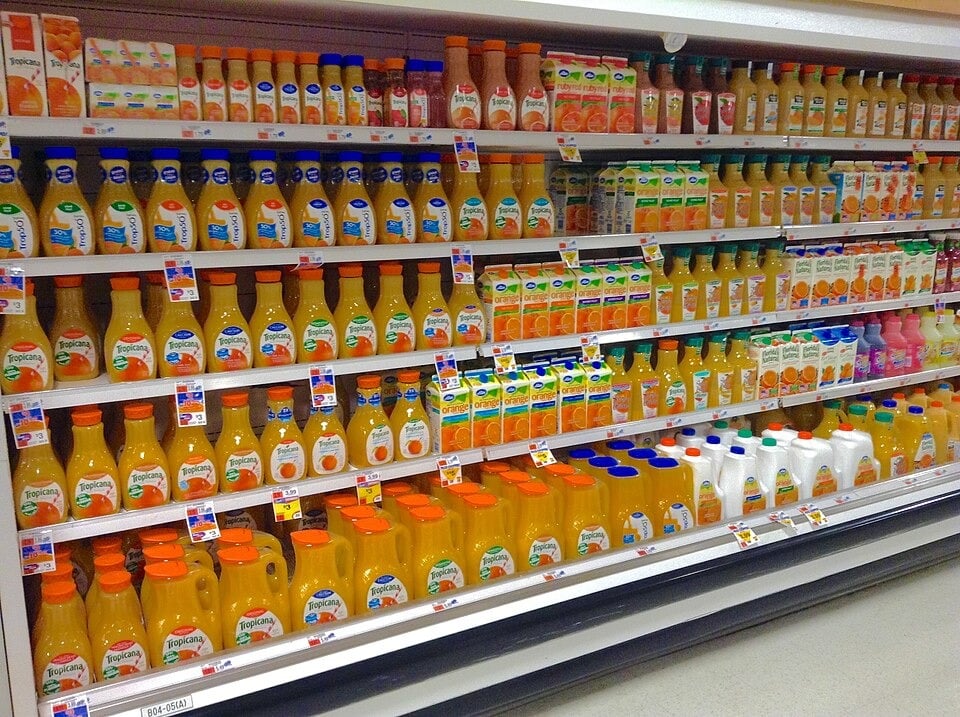 Supermarket refrigerated shelf filled with various brands and types of orange juice and other juice cartons and bottles, organized in rows.