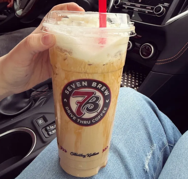 A hand holds a large iced coffee with foam in a clear cup featuring the Seven Brew Drive Thru Coffee logo, inside a car—reminding you of modern coffee houses on the go. The person's blue jeans and car interior are visible.