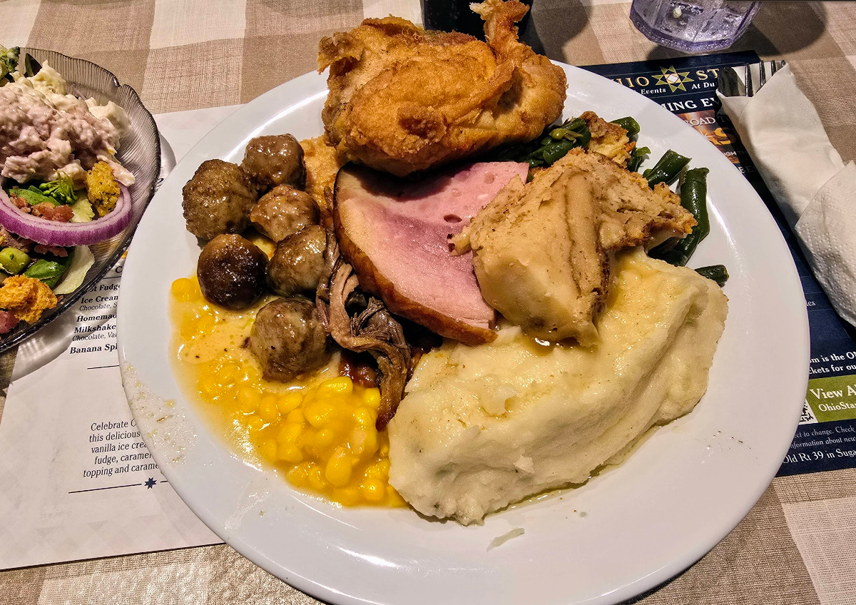 A plate of assorted foods—including mashed potatoes with gravy, corn, meatballs, sliced ham, fried chicken, green beans, and stuffing—shows the variety offered at a cheap all you can eat breakfast. A side salad with onion, lettuce, and egg is also visible.