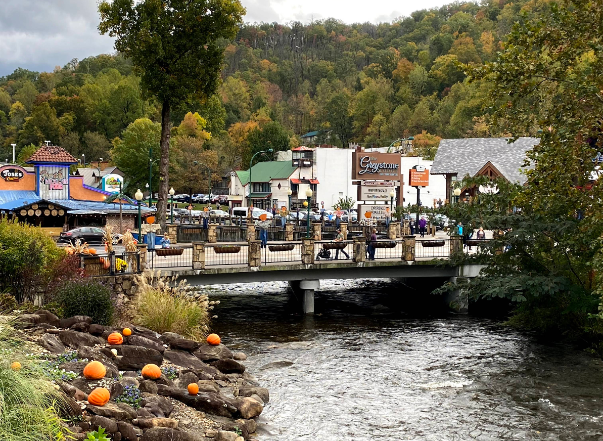 A bridge over a flowing creek in a small tourist town, with people walking, shops and signs in the background, and pumpkins scattered on the grassy bank near the water. Trees with autumn foliage cover the hills.