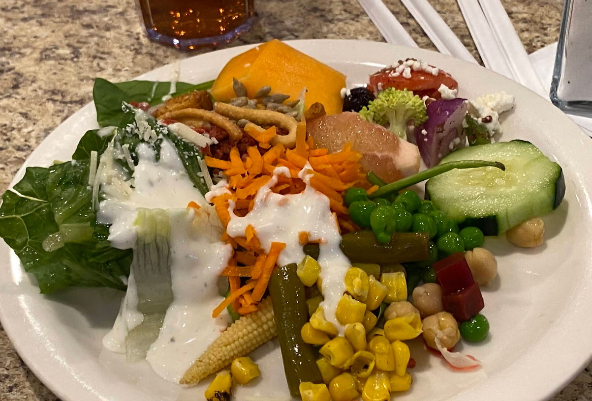 A plate of mixed salad with lettuce, shredded carrots, peas, corn, baby corn, cucumber, tomatoes, chickpeas, broccoli, and salad dressing—part of a cheap all you can eat breakfast—served with a cantaloupe slice and a glass in the background.