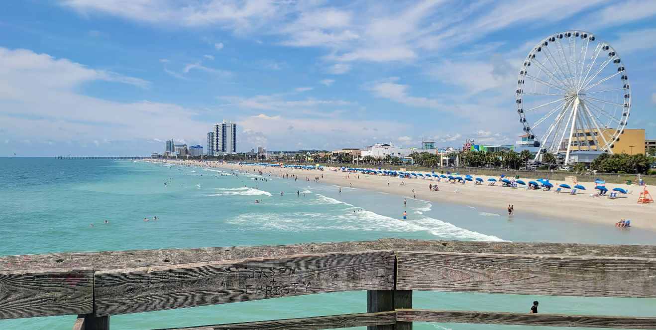 View of a beach with turquoise water, people swimming, a line of blue umbrellas, tall buildings, and a large Ferris wheel under a partly cloudy sky, seen from a wooden railing—capturing the lively atmosphere often found in popular tourist towns.