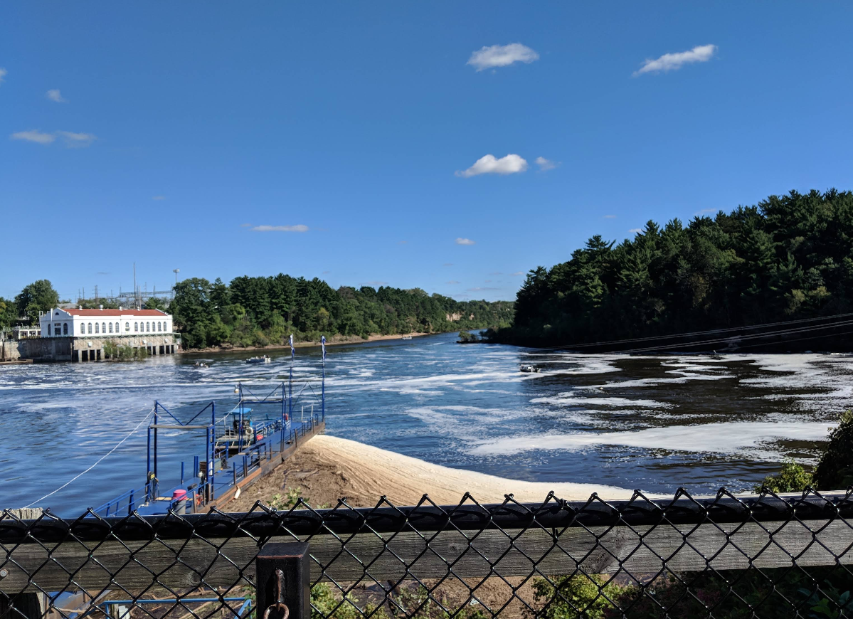 A river flows between forested banks under a blue sky with scattered clouds. A building is visible on the left, and a fenced observation area overlooks the water—a tranquil spot often found in charming tourist towns.