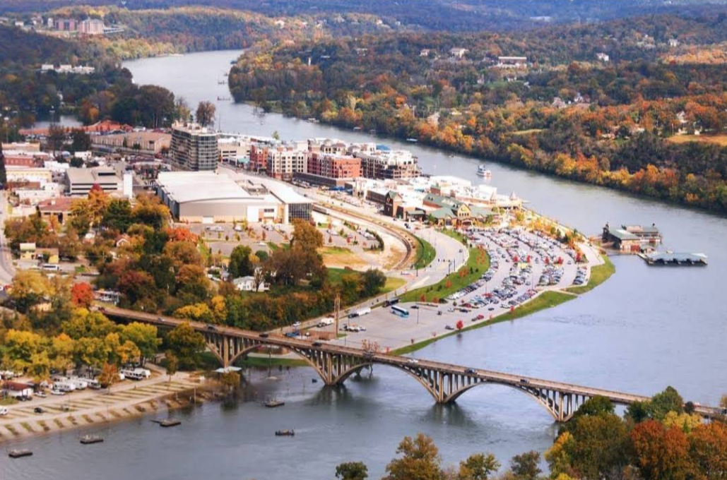 Aerial view of a bridge over a river in one of the region's charming tourist towns, with a city, parking lots, and boats nearby, surrounded by trees showing autumn colors.