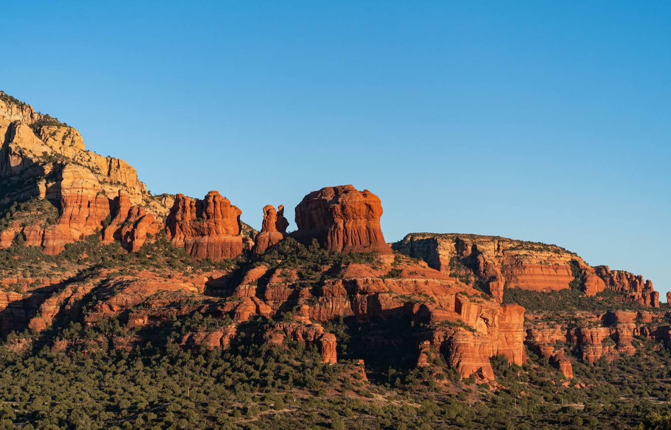Red rock formations in Sedona, Arizona, are brightly lit by the sun under a clear blue sky, with green vegetation at the base of the rocks—a stunning scene that has made Sedona one of the most popular tourist towns in the region.