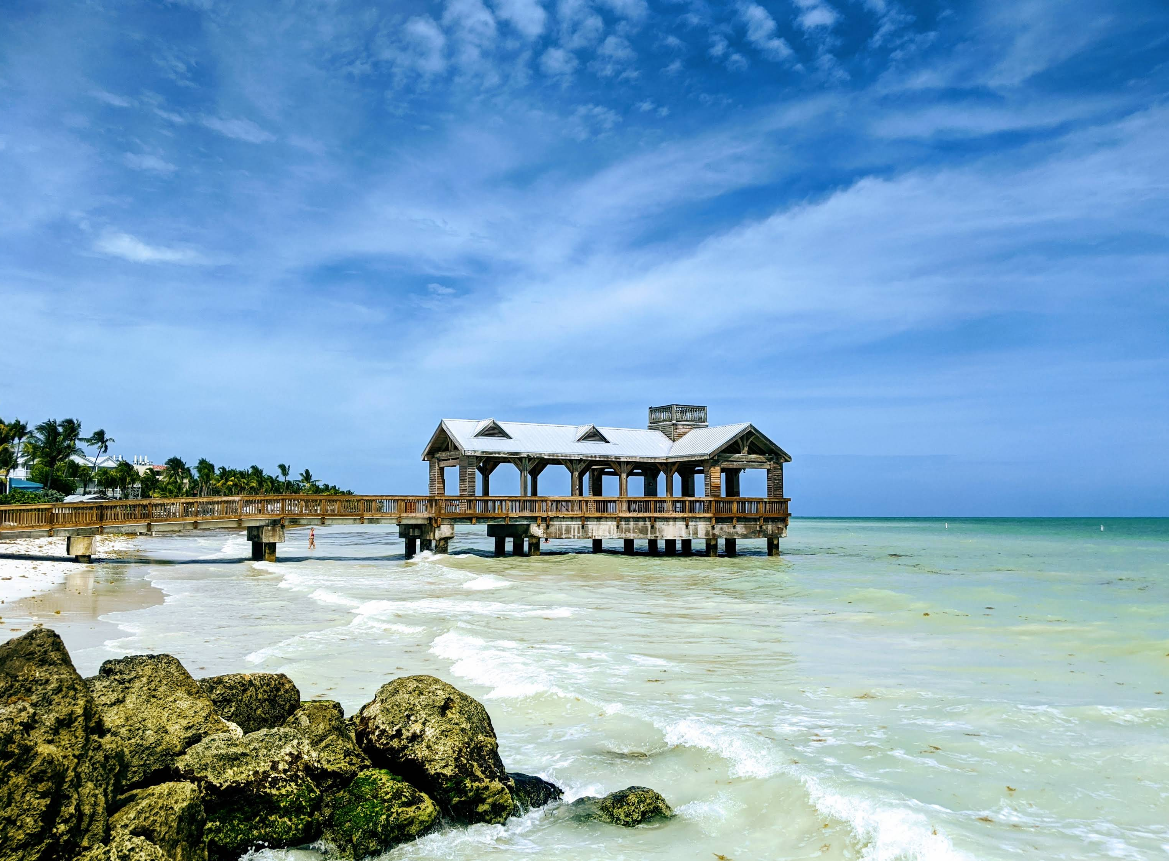 A wooden pier with a covered structure extends over turquoise water under a partly cloudy sky, with waves, rocks in the foreground, and palm trees lining the beach in the background—capturing the inviting charm often found in vibrant tourist towns.