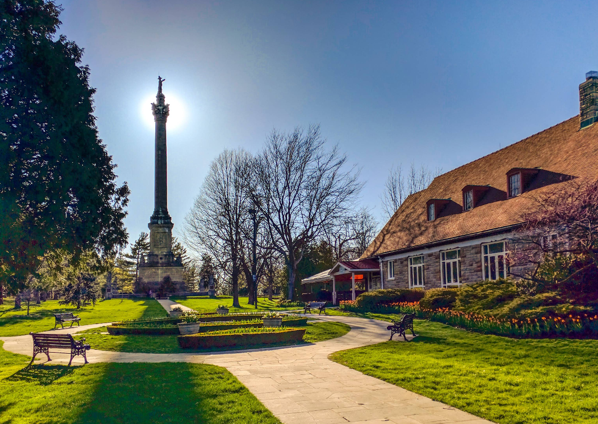 A tall monument stands in a sunlit park with a historic stone building and flowerbeds nearby—a tranquil spot often found in charming tourist towns. Paths, benches, and trees surround the peaceful scene on a clear day.