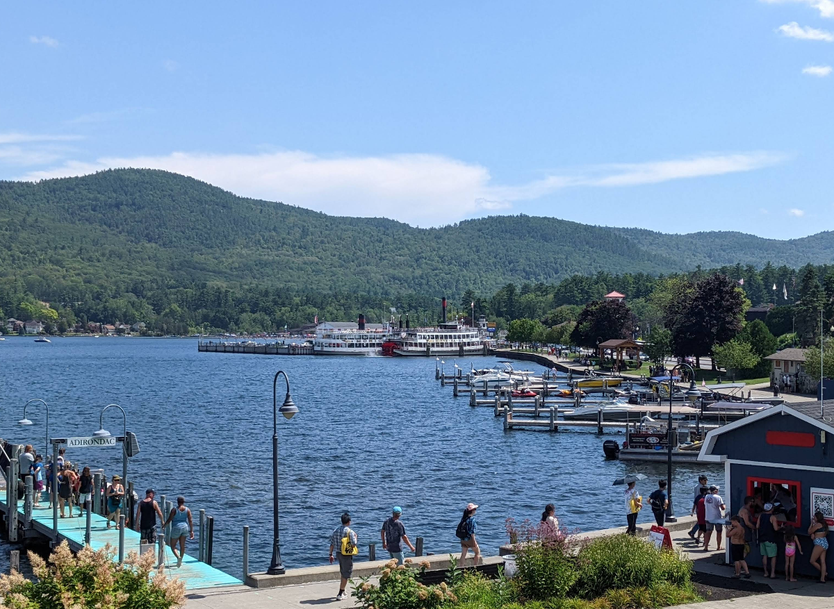 A lakeside marina with docks, boats, and a crowd of people captures the lively spirit often found in popular tourist towns. A large white steamboat is docked at the shore, while green hills and trees surround the lake under a blue sky with scattered clouds.