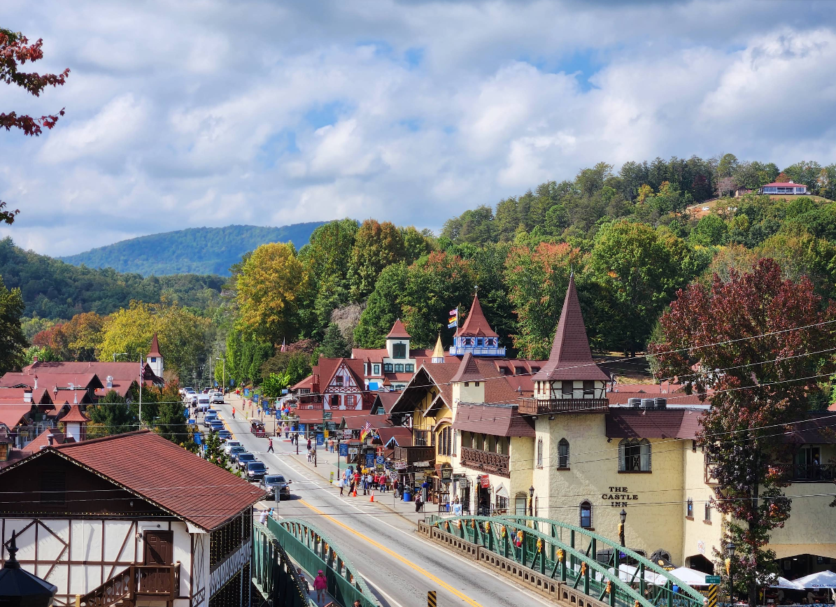 A scenic view of a small Bavarian-style tourist town nestled among green hills, with charming buildings, a busy street, people walking, and cars parked along the roadside under a partly cloudy sky.