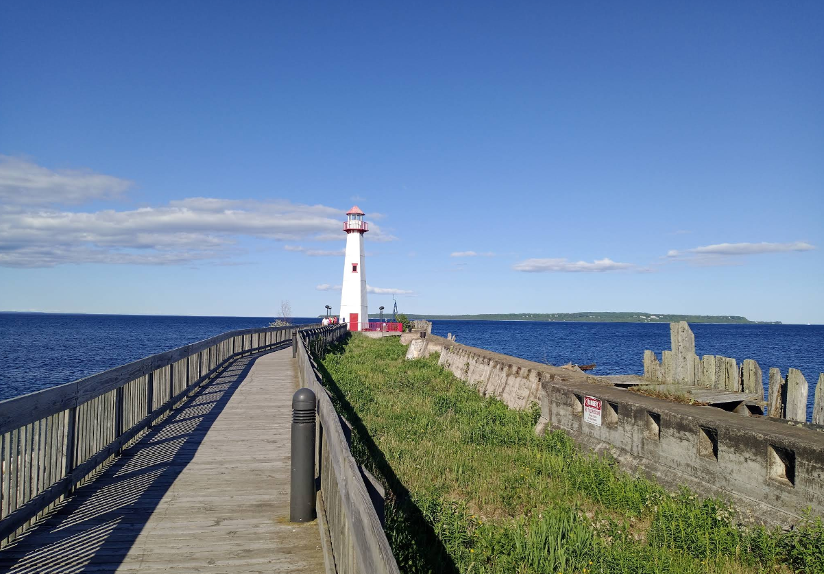 A white lighthouse with a red top stands at the end of a wooden pier by the blue sea, with grassy areas and old stone remains under a clear sky—a picturesque scene often found in charming tourist towns.
