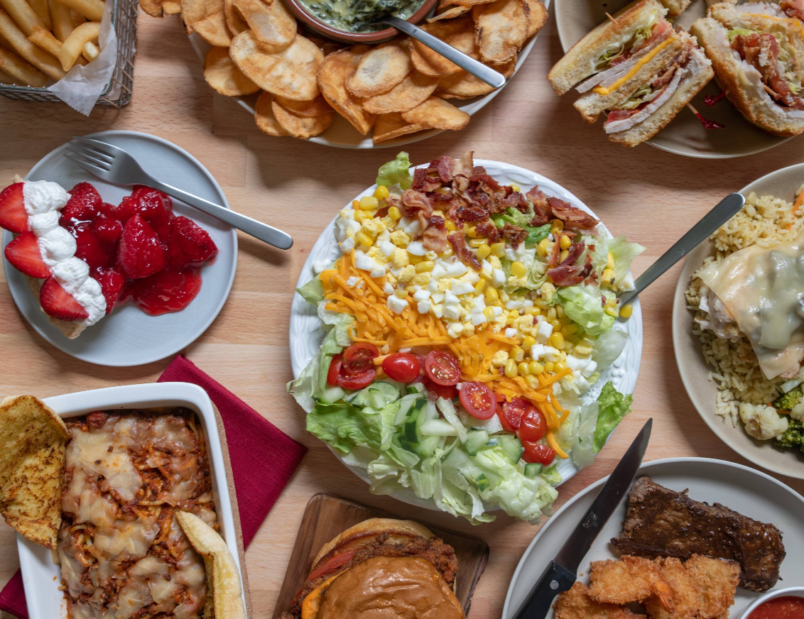 A table filled with various foods, like a hearty salad with egg, bacon, cheese, and tomatoes, potato chips, burger, fried chicken, lasagna, strawberry dessert—just what you’d expect at a cheap all you can eat breakfast buffet.