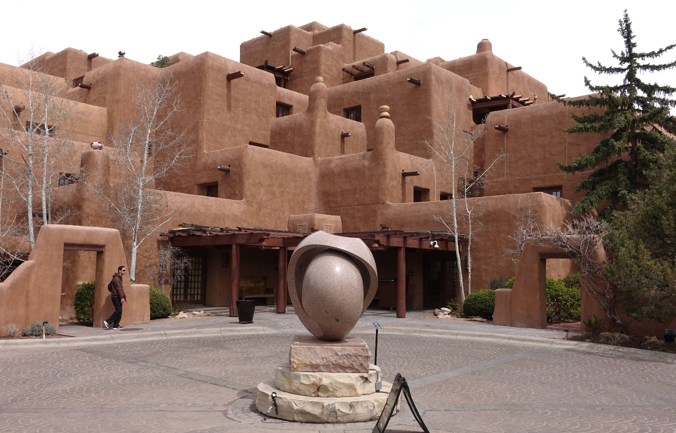 Brown adobe-style building with wooden accents, leafless trees, and a round stone sculpture in the courtyard. In the style of many charming tourist towns, a person stands on the left near an archway. Pine tree on the right.