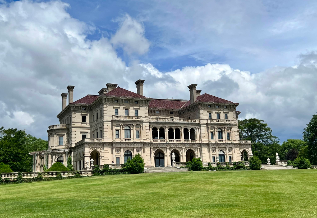 A grand, ornate mansion with a red roof sits on a large, manicured lawn under a partly cloudy sky—just the kind of architectural marvel you might find in charming tourist towns. The building features arches, columns, and elaborate stonework, while trees and statues are visible in the background.