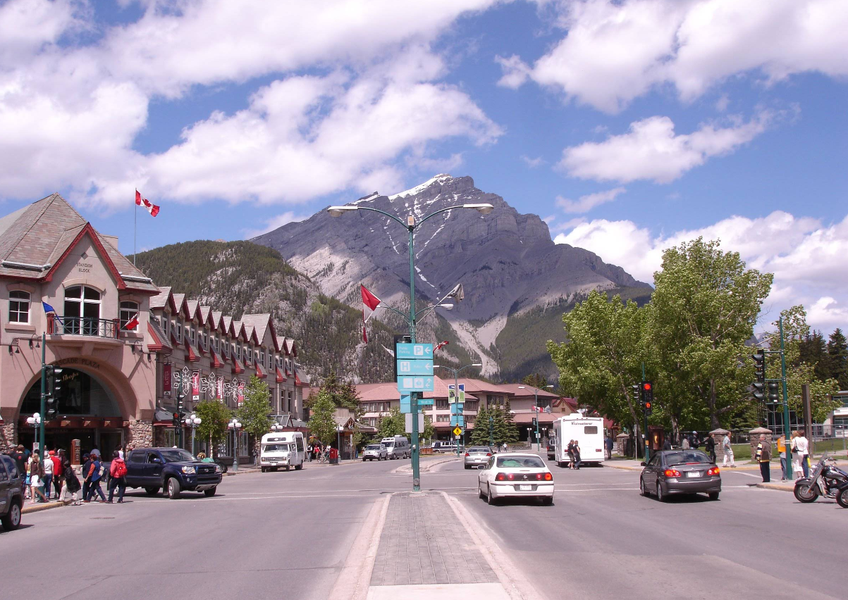 A busy street in one of the region’s charming tourist towns, with cars, people, Canadian flags, and a row of buildings, set against the dramatic backdrop of a large, rocky mountain rising under a partly cloudy sky.