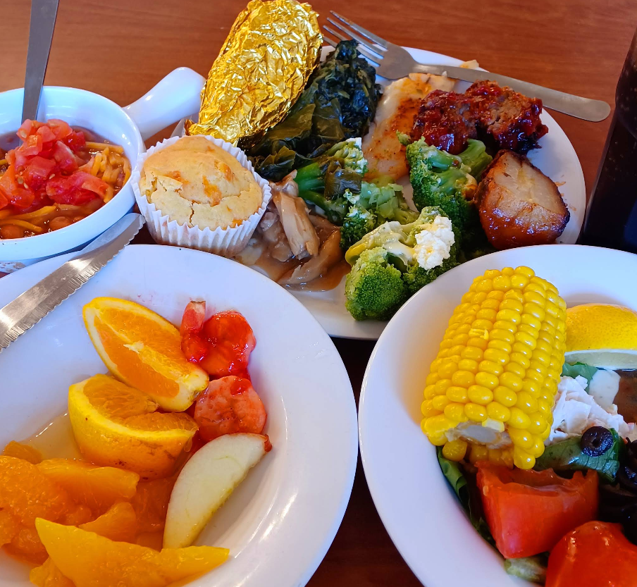 A colorful meal inspired by a cheap all you can eat breakfast, featuring plates of cornbread, foil-baked potato, chicken, broccoli, greens, roasted potatoes, salad with corn on the cob and tomatoes, plus a fruit bowl with orange slices and peaches.