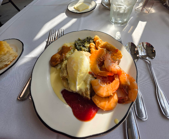 A plate of food featuring shrimp, a biscuit, collard greens, smothered chicken topped with creamy sauce, and cranberry sauce, set on a white tablecloth—perfect for a cheap all you can eat breakfast with buttered bread and a glass of water.