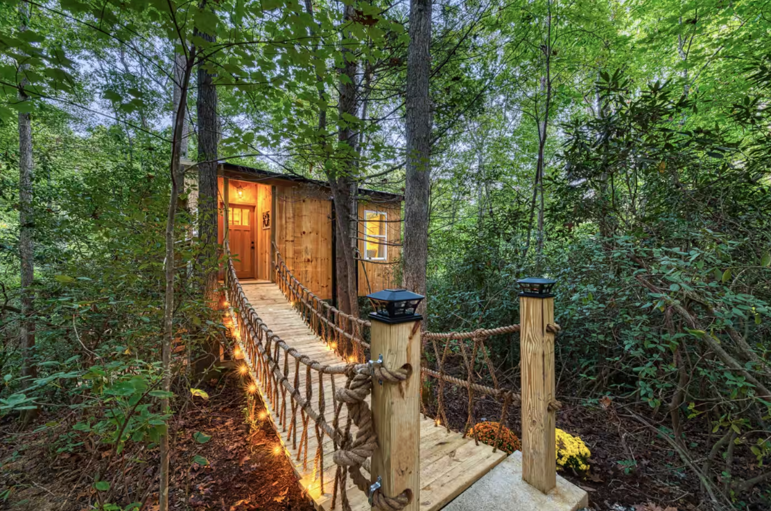 A small wooden treehouse with a rope bridge, surrounded by lush green trees. Warm lights line the bridge, and the treehouse door is open, revealing a cozy interior glow.