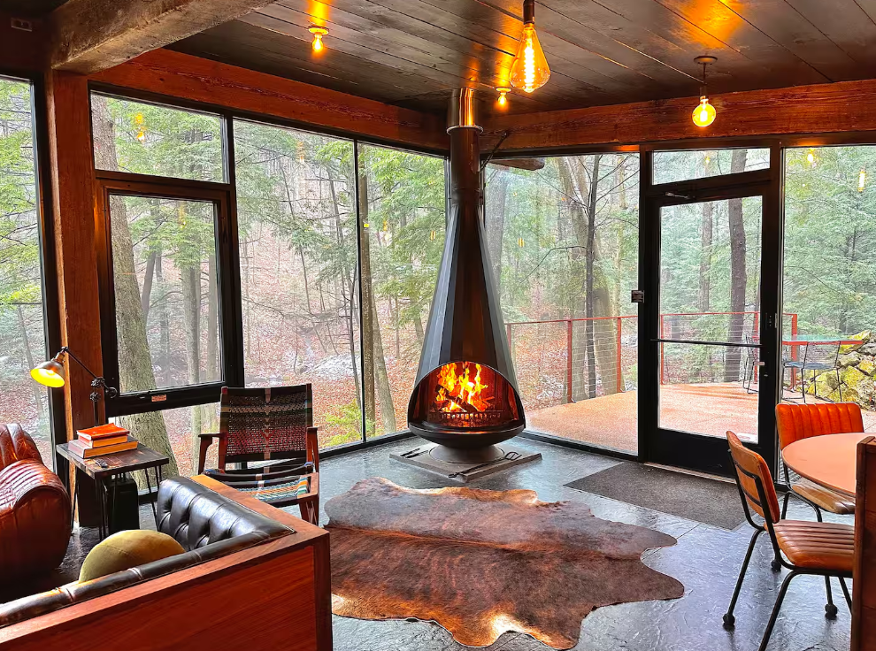 A cozy living room with floor-to-ceiling windows, a modern hanging fireplace, leather and wooden furniture, a cowhide rug, and forest views outside. Warm light bulbs hang from the ceiling.