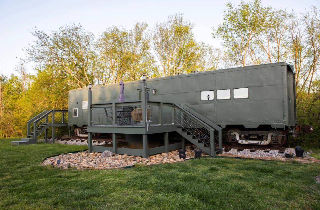 An olive-green train car sits on tracks in a grassy yard, surrounded by trees. A raised wooden deck with patio furniture and railing is attached, with a small stone area and stairs leading to the entrance.