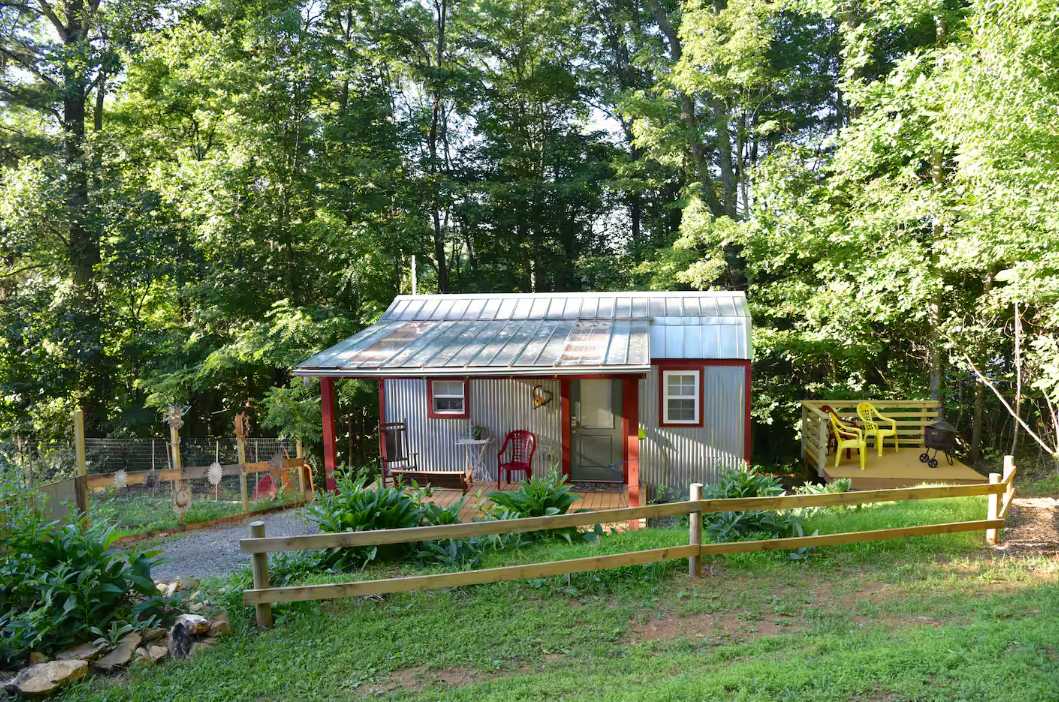 A small red and gray cabin with a metal roof sits in a wooded area, surrounded by greenery and a wooden fence. There’s a gravel path, red chairs on the porch, and a yellow bench on a deck to the right.