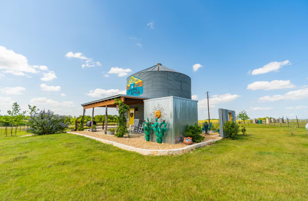 A small, round metal building with a porch sits on a grassy field under a blue sky with scattered clouds. There are colorful decorations, plants, and a cactus sculpture near the entrance.