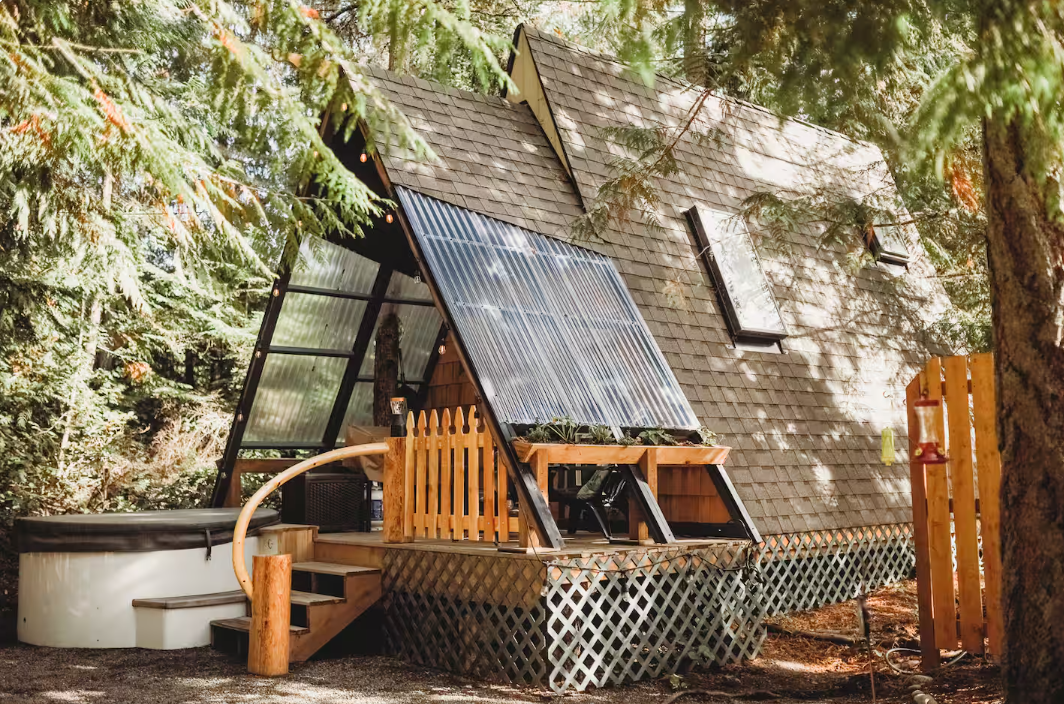 A cozy A-frame cabin with a wooden deck and solar panels is nestled among tall trees in a forest setting. A small picket fence and outdoor seating area are visible, blending with the natural surroundings.