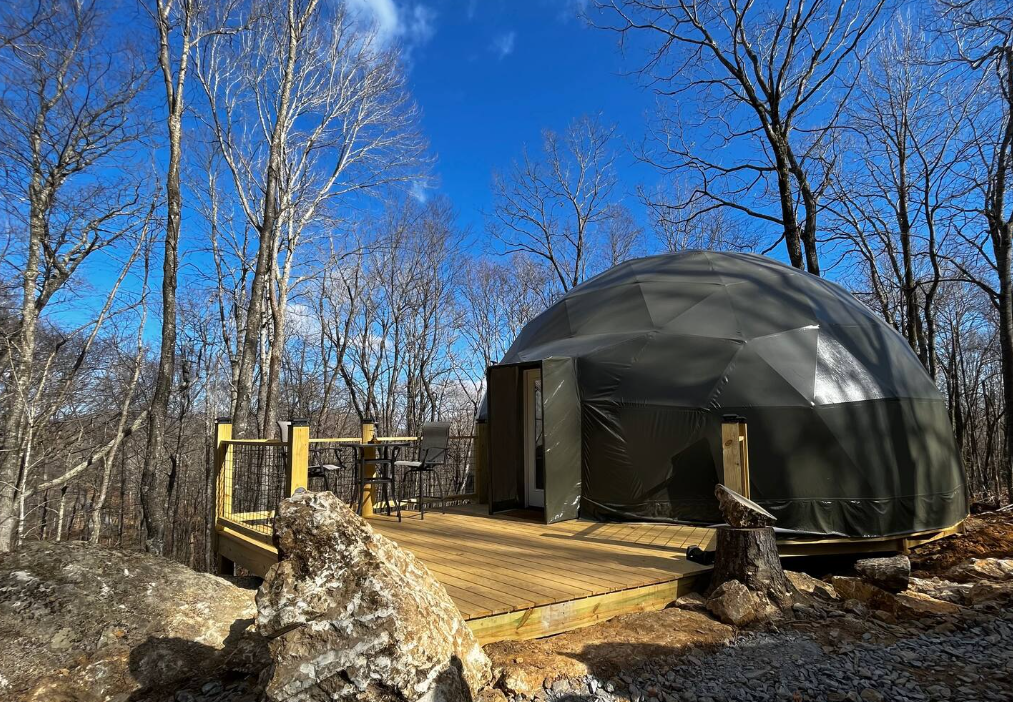 A dome-shaped tent sits on a wooden deck in a forested area with leafless trees, a table and chairs nearby, and blue sky above.