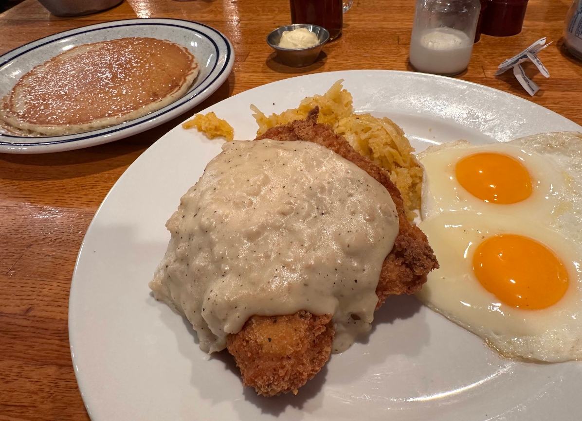 A plate with chicken fried steak topped with gravy, scrambled eggs, and two sunny side up eggs, plus a side of pancakes and syrup in the background—all part of a cheap all you can eat breakfast on a wooden table.