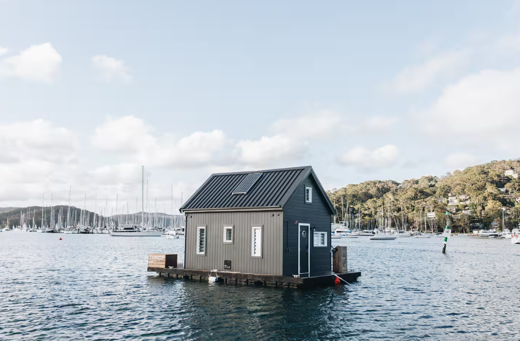 A small grey house floats on calm water, surrounded by boats and distant hills under a partly cloudy sky.