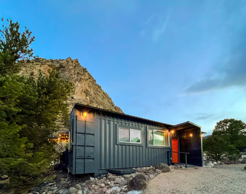 A small black house made from a shipping container sits on rocky ground near green trees and a mountain, lit warmly at dusk under a blue sky.