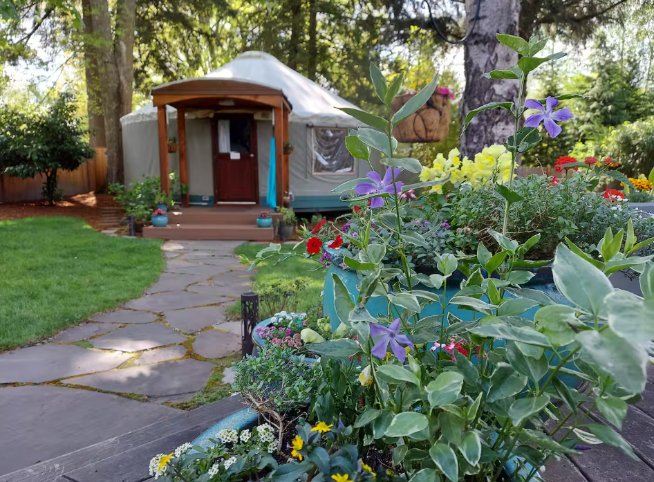 A lush garden with colorful flowers and stone path leads to a round yurt surrounded by trees. The yurt has a small porch and wooden door, blending with the natural, peaceful setting.