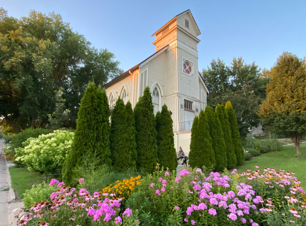 A charming light-colored church with a tall tower stands behind neatly trimmed evergreens and colorful blooming flowers, surrounded by lush green trees and grass on a sunny day.