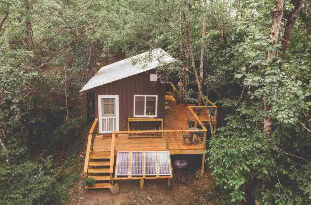 A small brown cabin with a metal roof sits in a forest. It has a wooden deck, outdoor seating, and solar panels on the ground in front. Trees surround the cabin, creating a peaceful, secluded atmosphere.