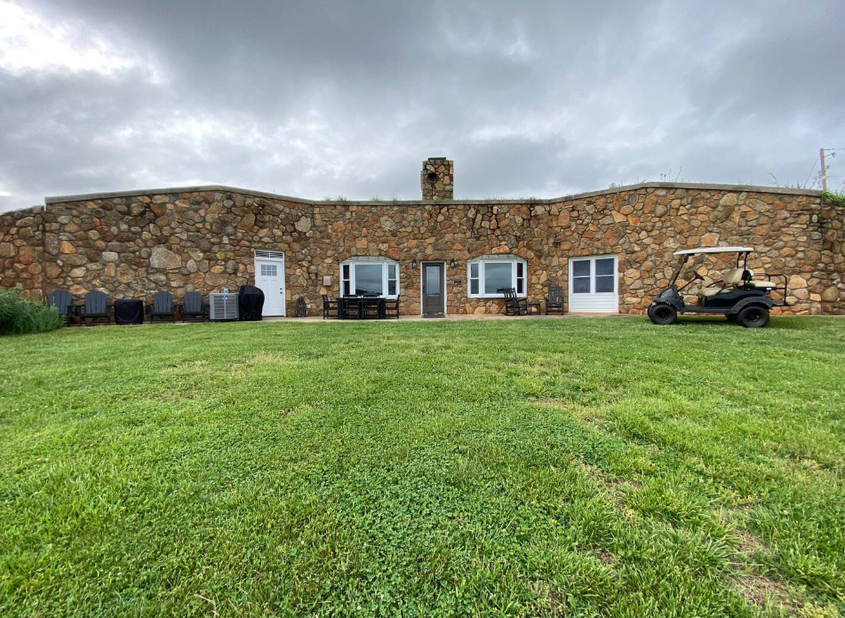 A stone building with multiple doors and windows is set under a cloudy sky. Black Adirondack chairs are on the left, and a golf cart is parked on the right, all on a large, green lawn.