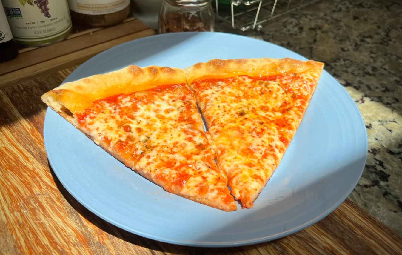 Two slices of cheese pizza with golden, bubbly cheese and tomato sauce sit on a light blue plate atop a wooden surface in a sunlit kitchen, making for one of the classic, comforting meals.