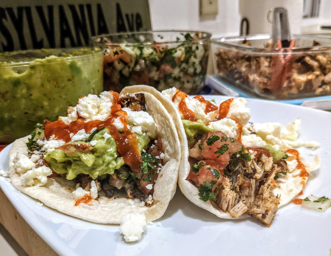 Two tacos filled with shredded meat, guacamole, salsa, queso fresco, and cilantro sit on a white plate—making for satisfying meals—with containers of guacamole, salsa, and shredded meat in the background.