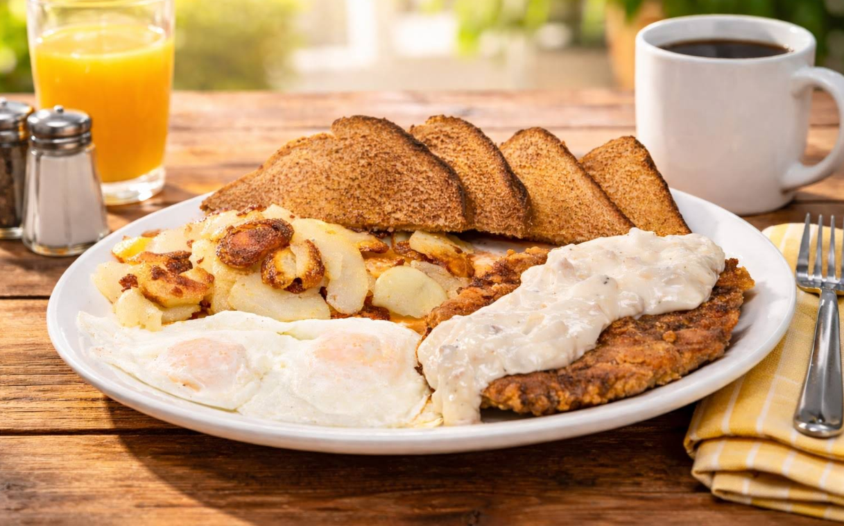 A breakfast plate with fried eggs, crispy potato slices, toast, and country fried steak topped with gravy—perfect for a cheap all you can eat breakfast. A cup of coffee and a glass of orange juice sit in the background on a wooden table.