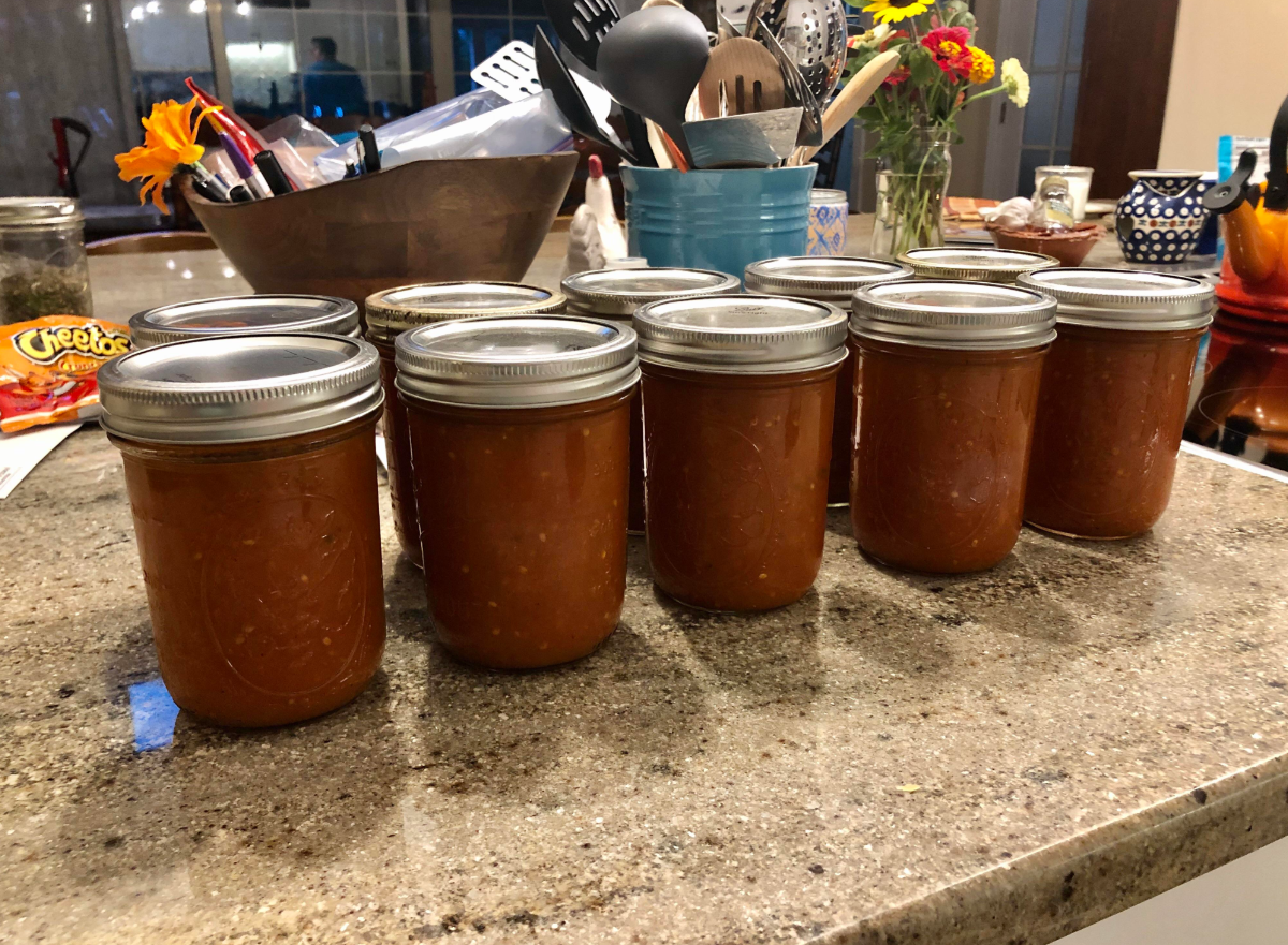 Nine glass jars filled with reddish-brown sauce are lined up on a kitchen counter, ready to enhance your future meals. Kitchen utensils, flowers, and Cheetos are visible in the background.