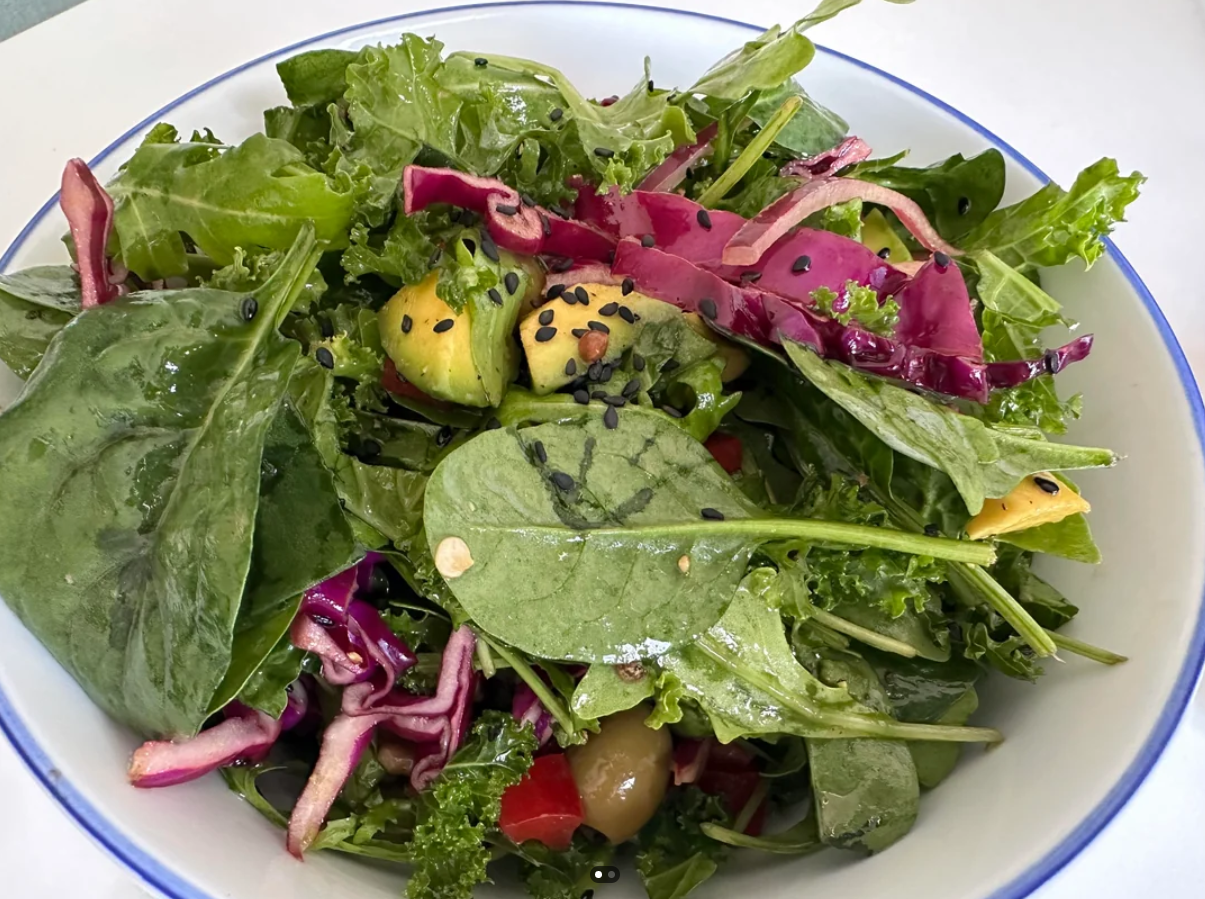 A close-up of a mixed green salad with spinach, kale, purple cabbage, sliced avocado, olives, and black sesame seeds in a white bowl with a blue rim—perfect for fresh and vibrant meals.