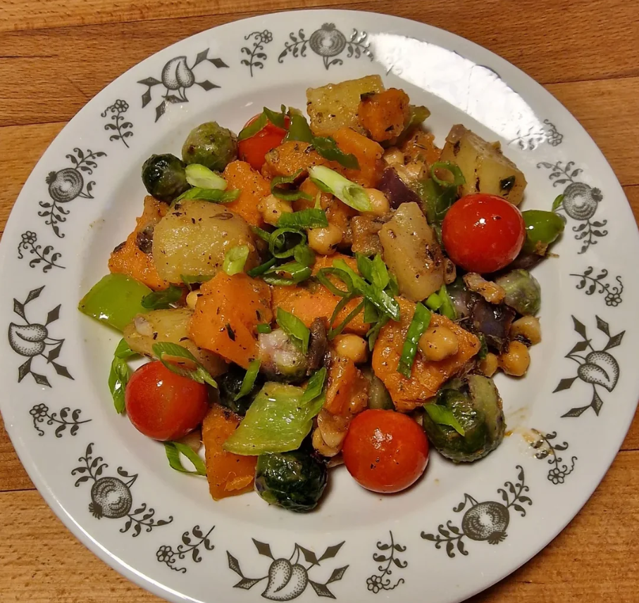 A plate of roasted vegetables, perfect for wholesome meals, including cherry tomatoes, Brussels sprouts, chickpeas, green onions, and diced squash, served on a white plate with a decorative floral pattern.