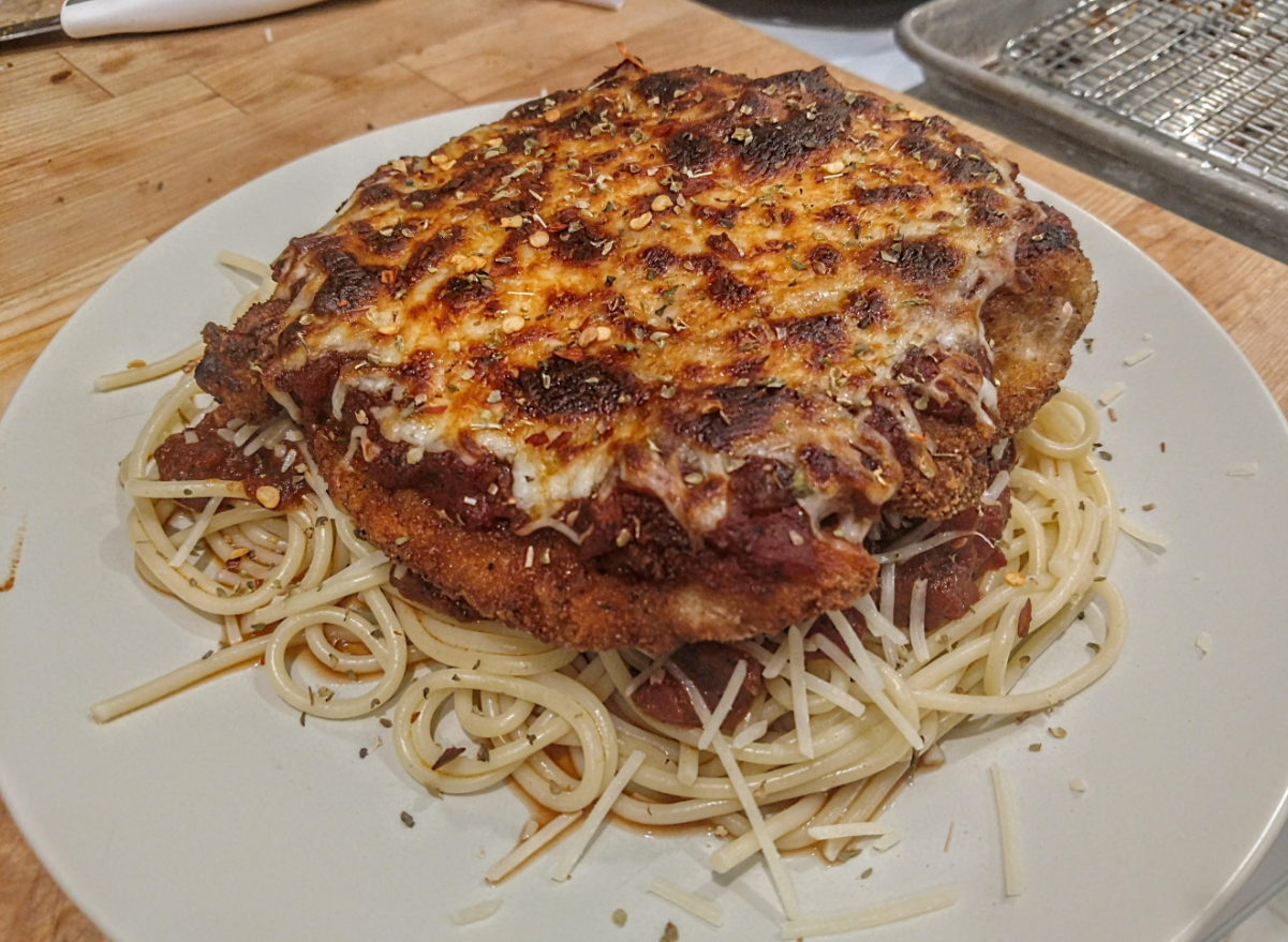 A plate of cooked spaghetti topped with a large breaded and baked chicken cutlet, covered in marinara sauce and melted cheese, makes for one of the most comforting meals, all beautifully arranged on a light-colored plate.
