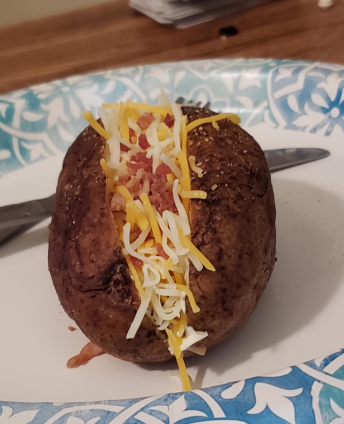 A baked potato topped with shredded cheese and bacon bits sits on a blue and white paper plate, making it a hearty addition to quick meals, with a fork in the background.