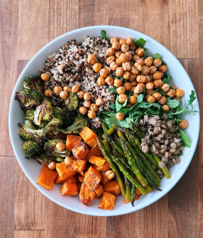 A white bowl filled with quinoa, roasted broccoli, sweet potatoes, asparagus, chickpeas, lentils, and arugula on a wooden surface makes for one of the most vibrant and nourishing meals.